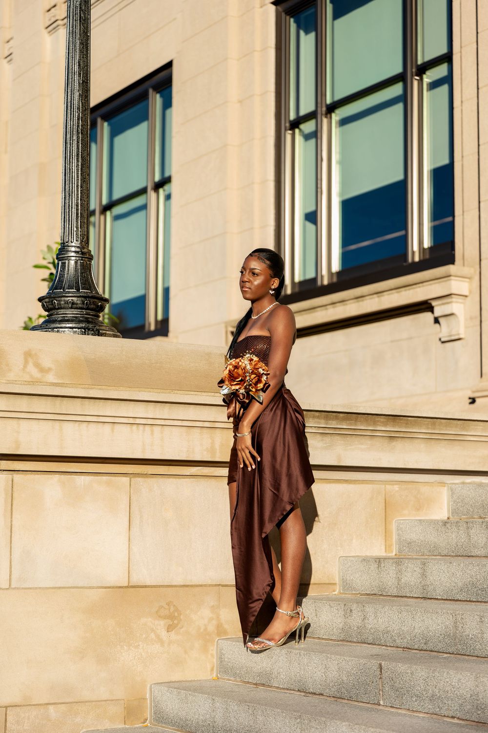Woman in a brown satin strapless dress and heeled sandals holds an autumn bouquet while standing on stone steps beside a sunlit building.