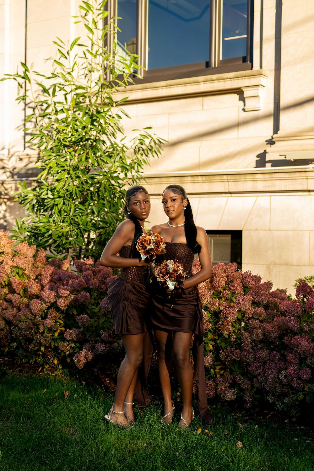 Two women in brown satin dresses hold autumn bouquets while posing outdoors beside blooming pink hydrangeas and a sunlit stone building.