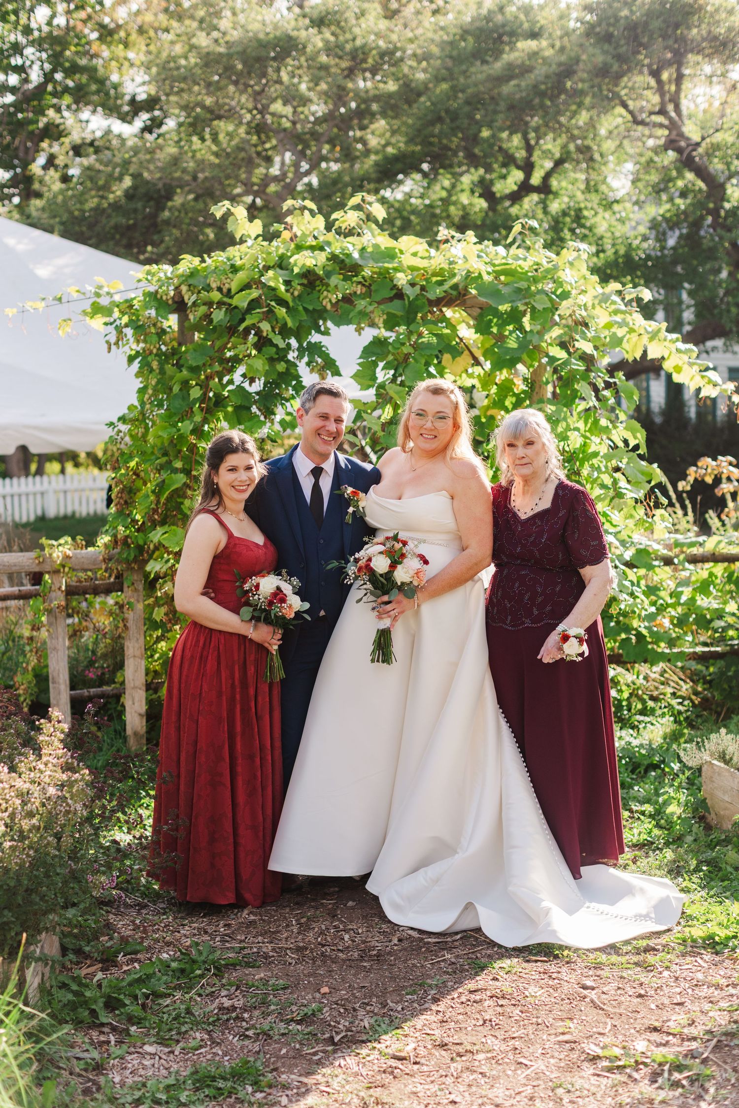 Bride in white gown and groom in navy suit pose with two women in burgundy dresses under a vine-covered garden arbor.
