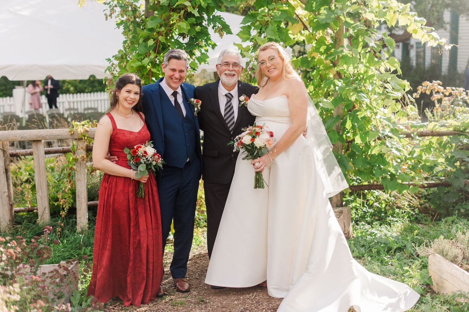 Bride in a white gown and veil poses with a bridesmaid in a red dress and two men in suits under a leafy garden arch.