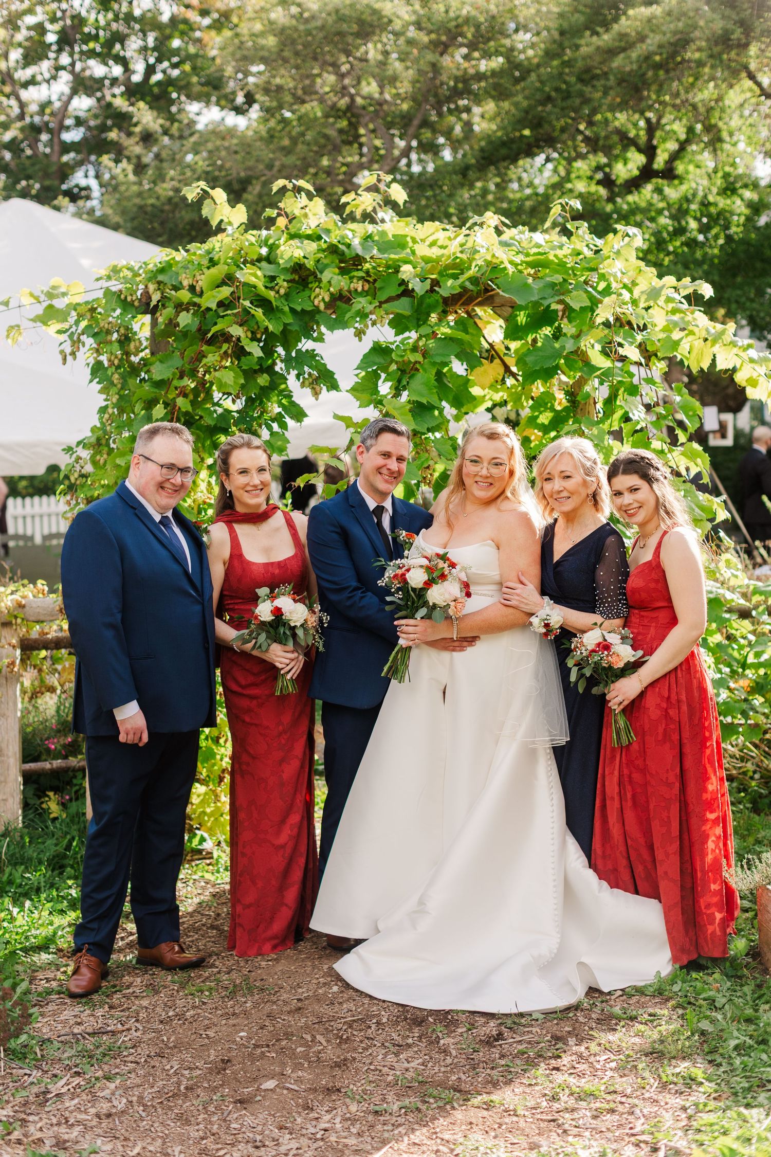 Bride in a white gown poses with the groom, two bridesmaids in red dresses, and a groomsman under a leafy garden arch at an outdoor wedding.