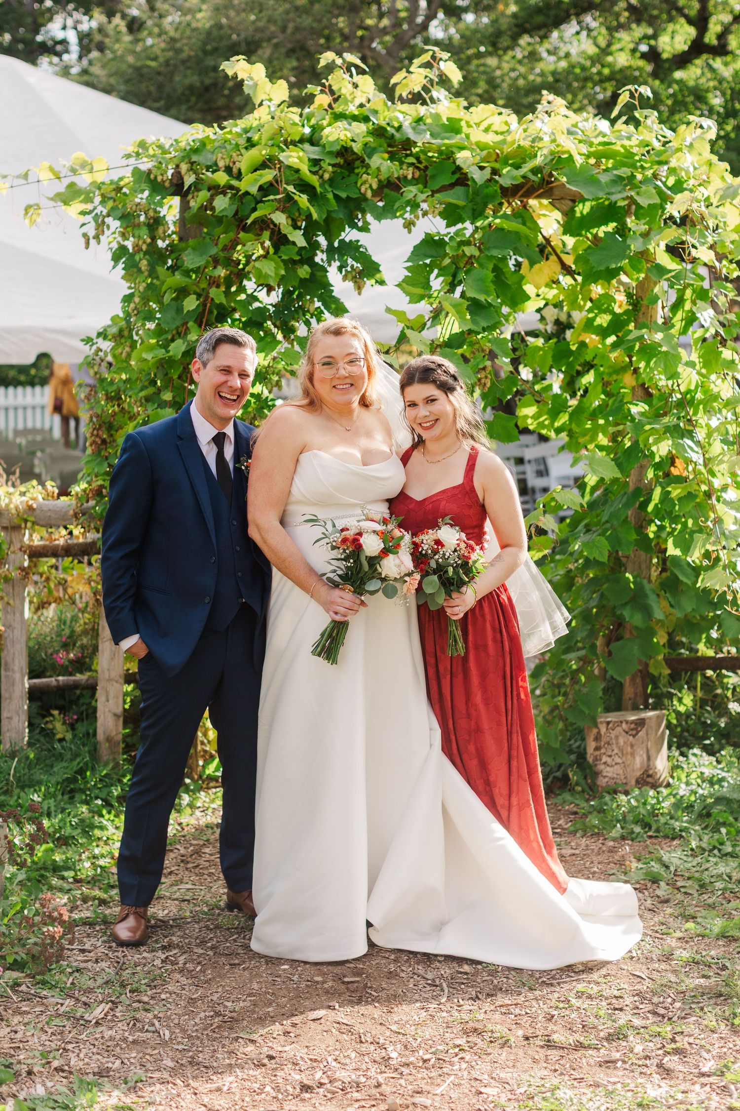 Smiling groom in a navy suit stands beside a bride in a white gown and a bridesmaid in red, holding bouquets under a leafy garden arbor.