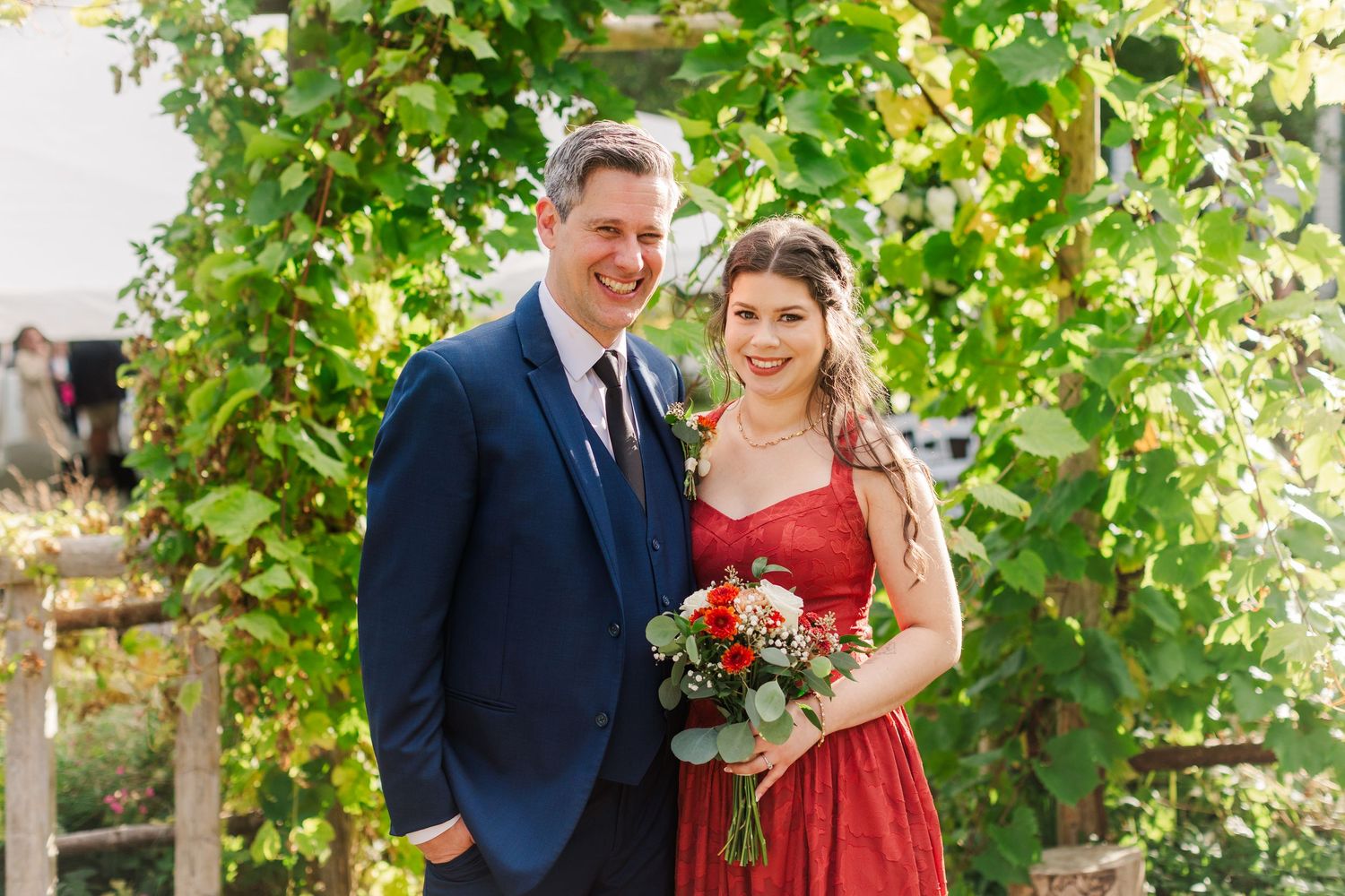 Smiling couple in a navy suit and red dress holding a bouquet of red and pink flowers beside a leafy green garden trellis.