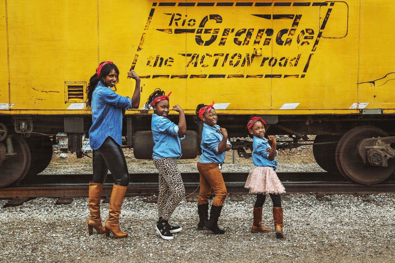 Four people in blue shirts and red bandanas pose in a line in front of a yellow Rio Grande train car on gravel tracks.