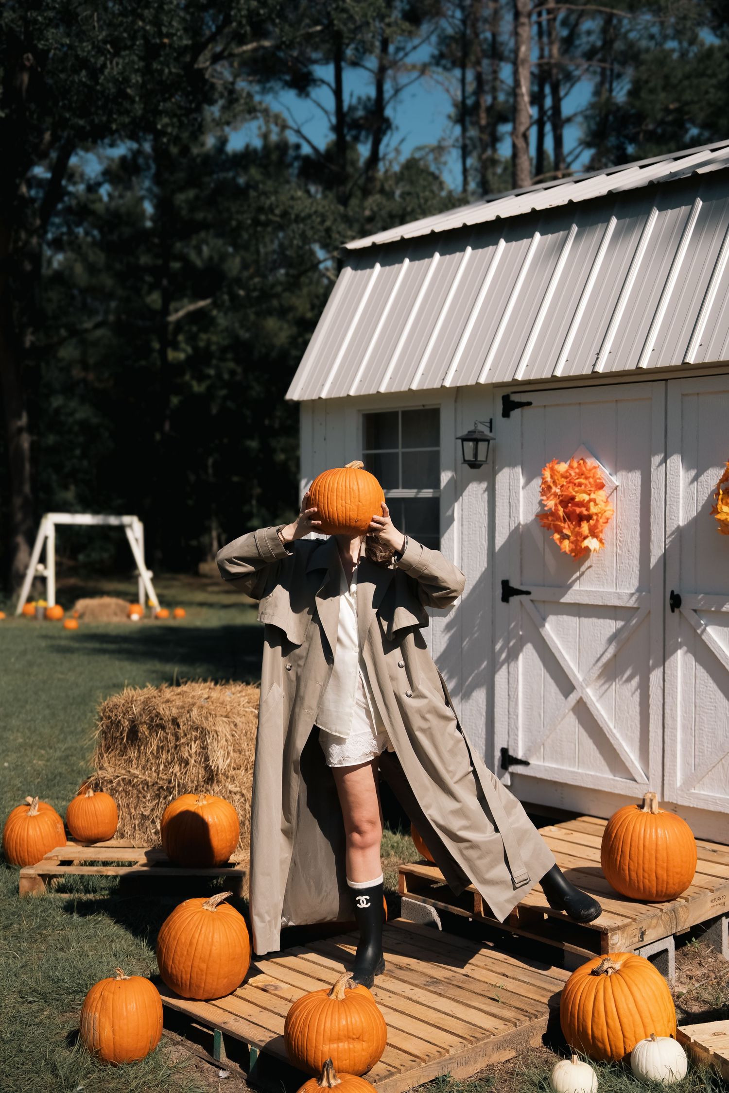 Person in a tan trench coat holds a pumpkin over their head beside a white shed with hay bales and pumpkins on a wooden platform.