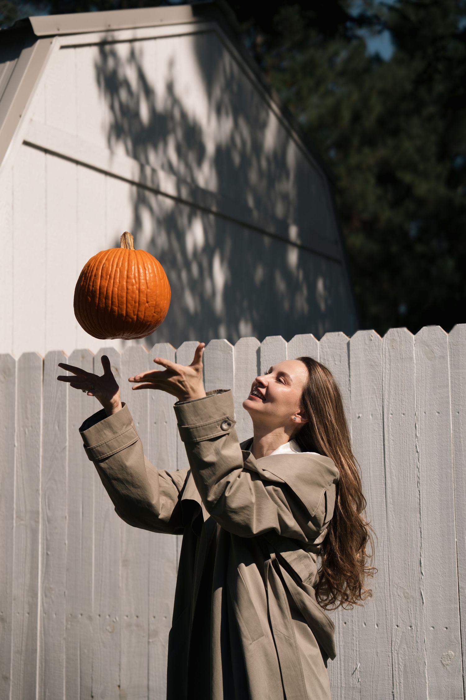 Smiling woman in a tan trench coat tosses an orange pumpkin into the air beside a white wooden fence on a sunny autumn day.