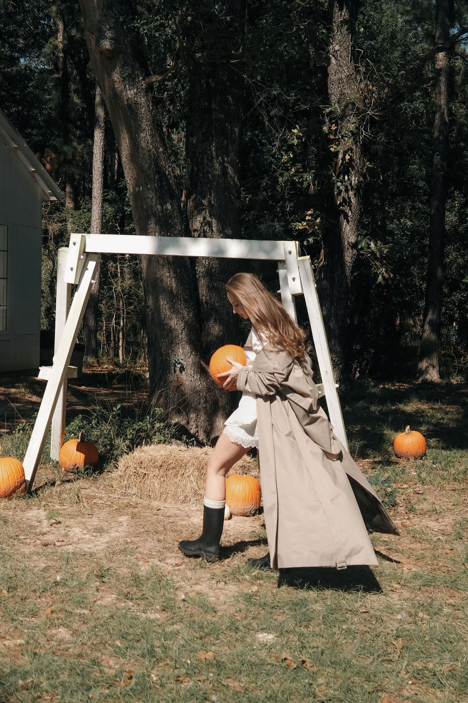 Woman in a tan trench coat and black rain boots carries a pumpkin past a wooden swing frame and hay bale in an autumn woodland yard.