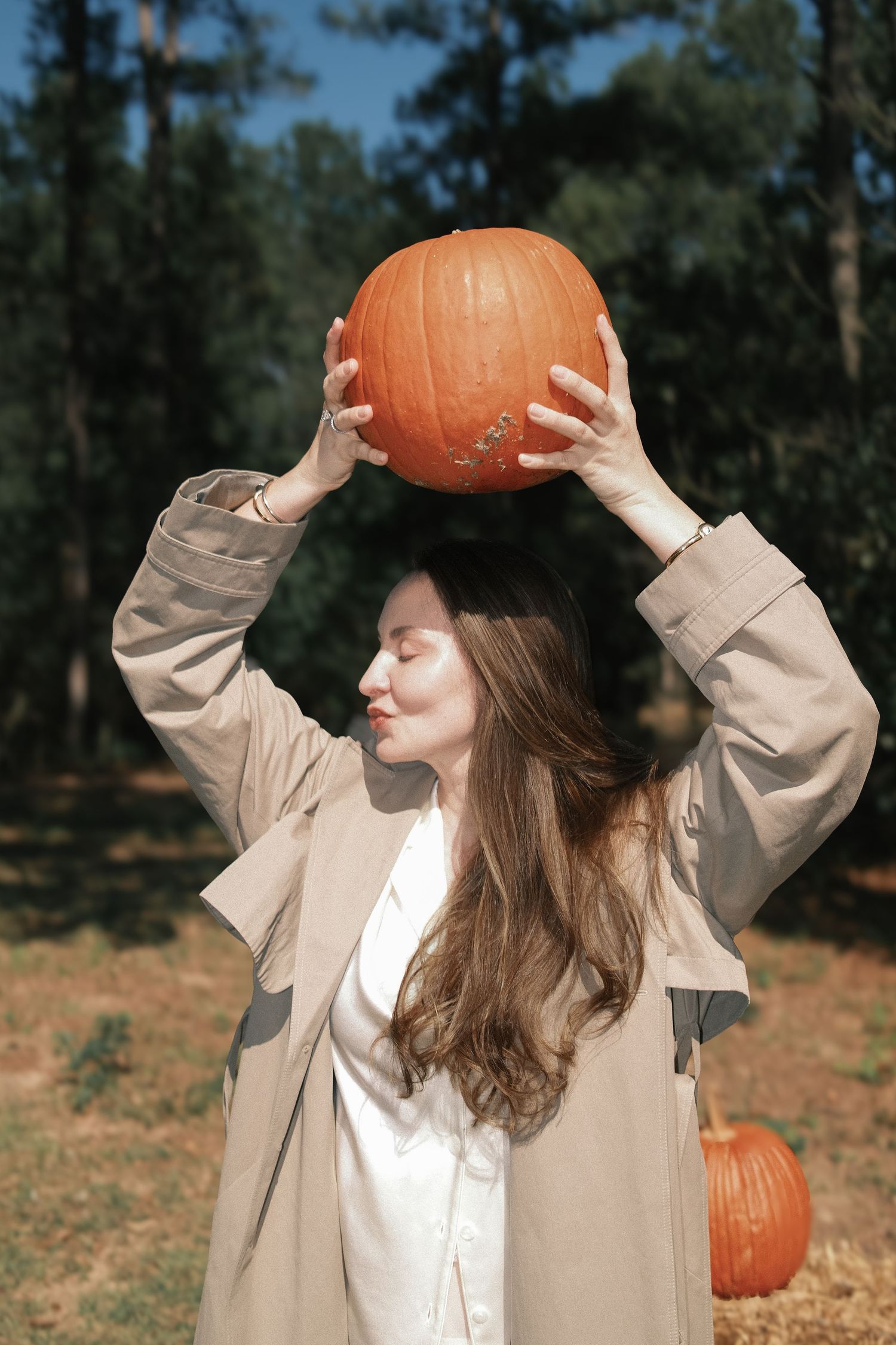 Woman in a beige trench coat holds a large orange pumpkin above her head in a sunny pine forest pumpkin patch.