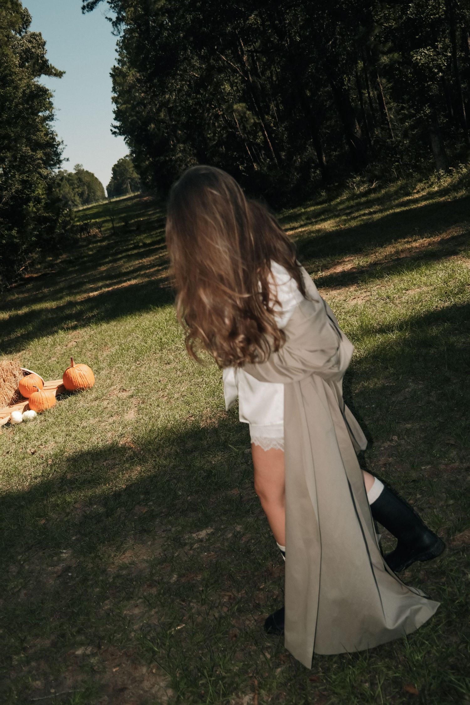 Woman with long wavy brown hair wearing a beige trench coat and black boots walks on a sunlit grassy field near pumpkins and trees.