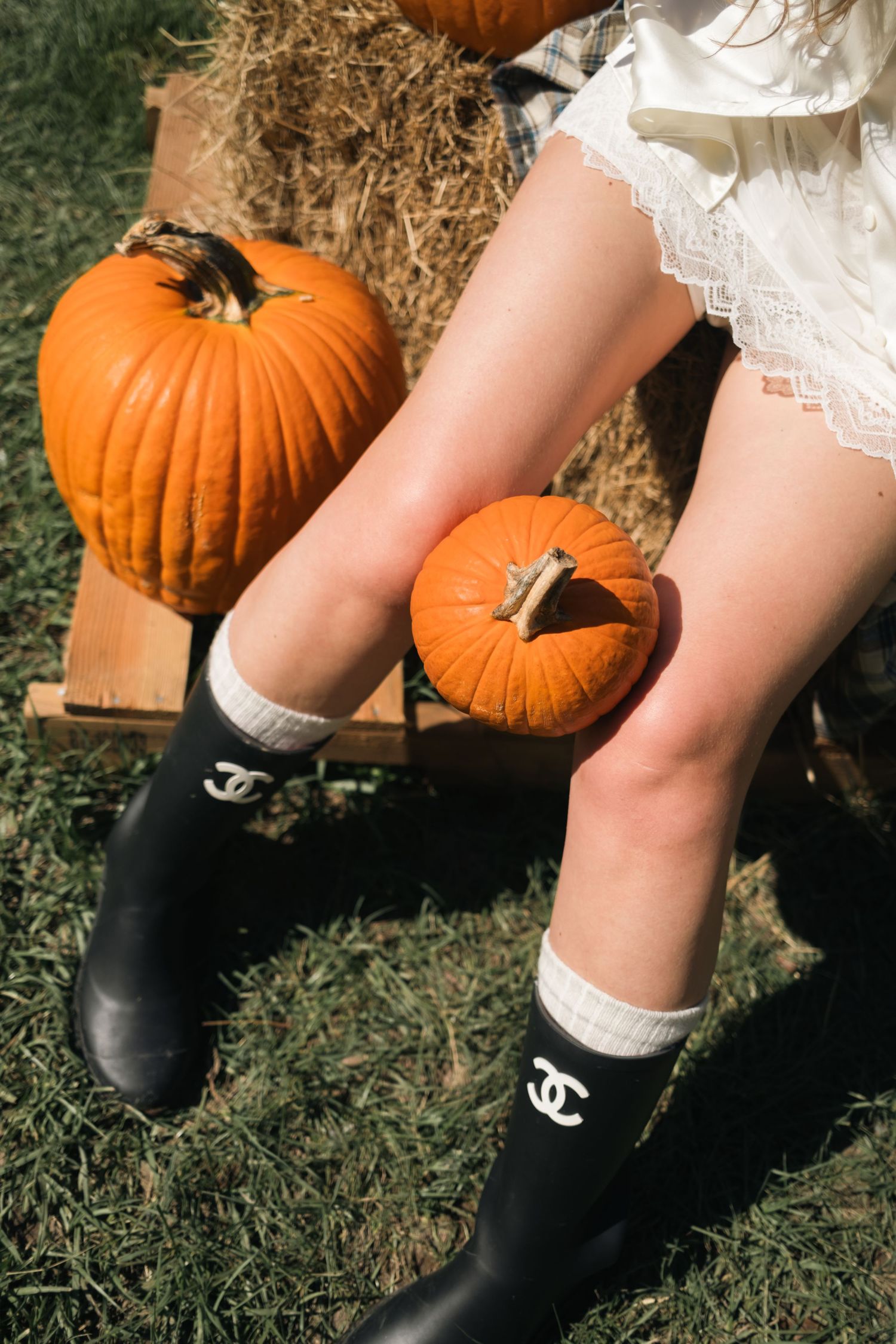 Woman in a white lace-trimmed dress and black rain boots with white socks sits on hay bales beside orange pumpkins on grass.