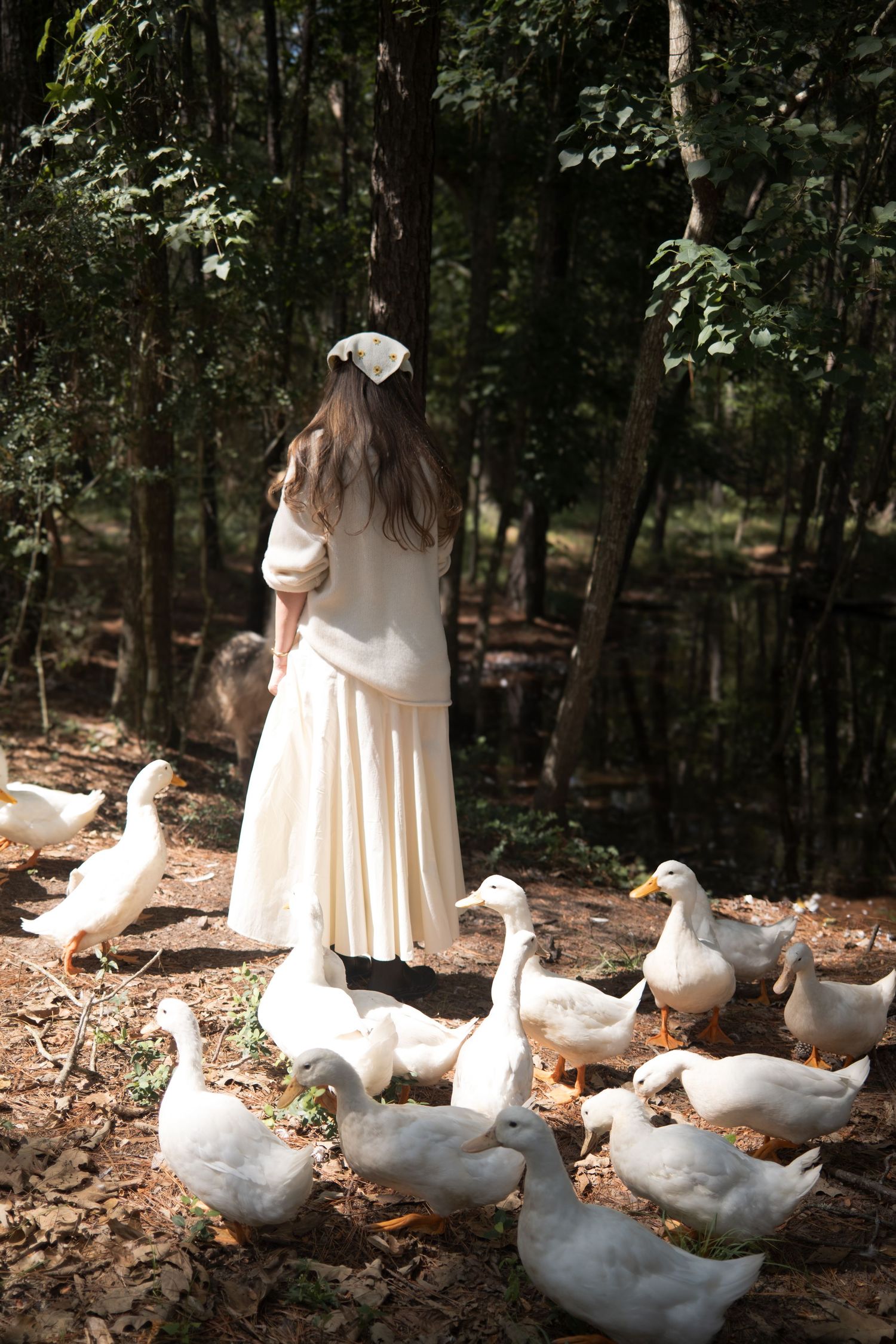 Woman in a white dress and hair bow seen from behind, surrounded by white ducks on a leaf-covered forest floor near a pond.