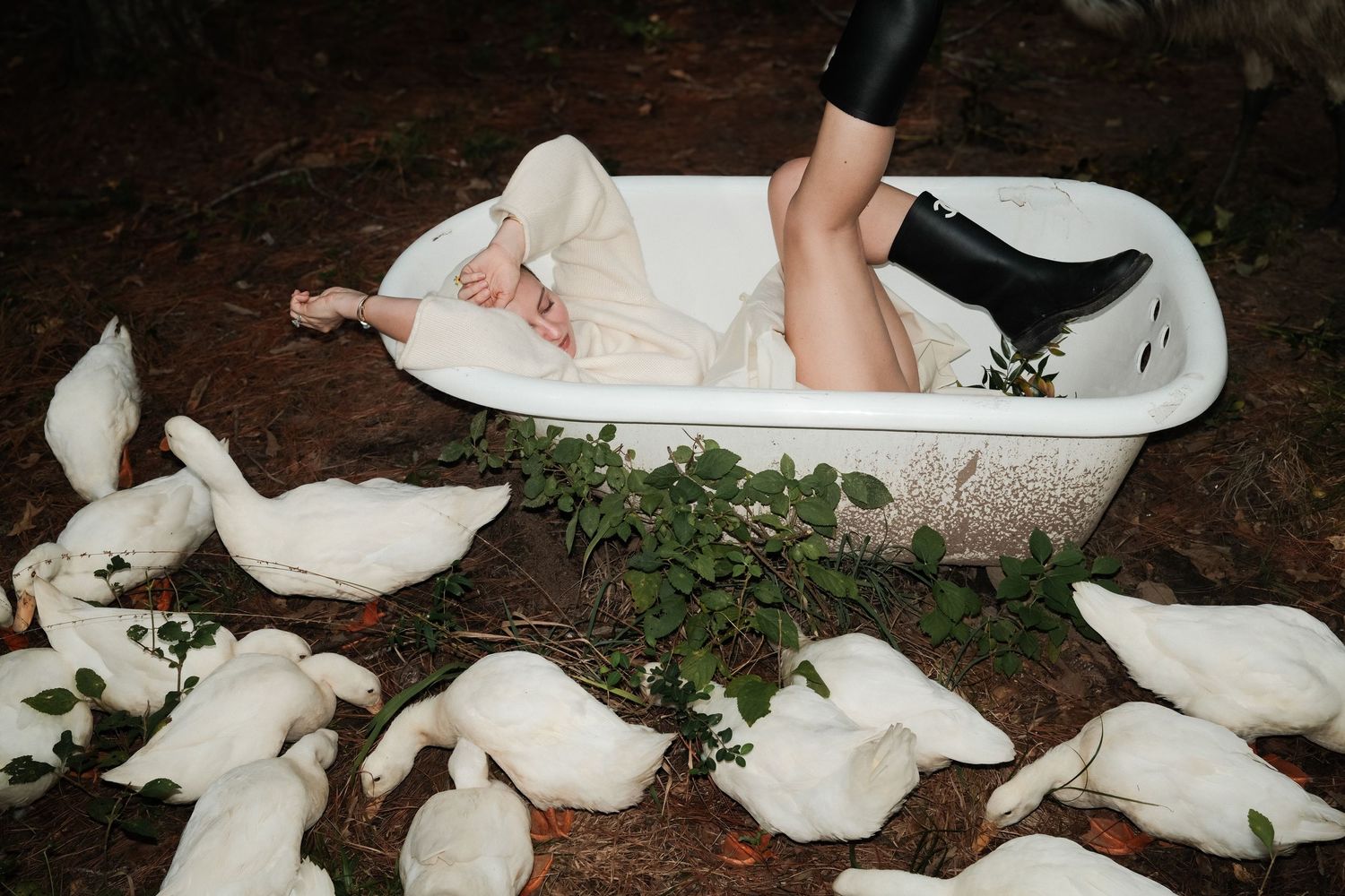 Woman in black boots reclines in a white bathtub on a forest floor surrounded by white ducks and green foliage.