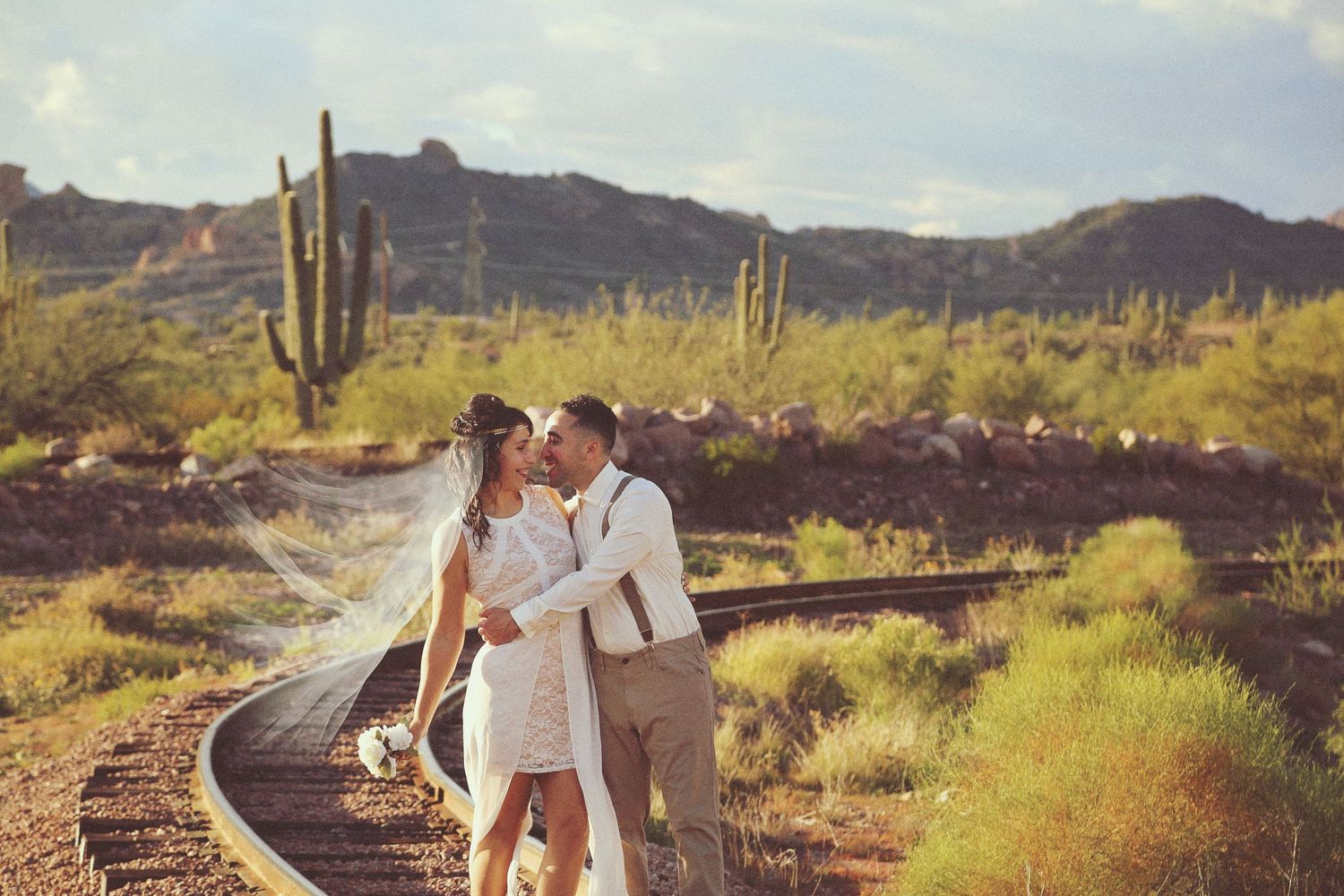 A bride and groom share a kiss on curved railroad tracks in the Arizona desert with saguaro cacti and mountains behind them.