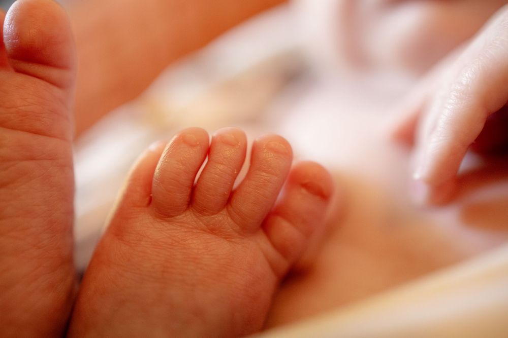 Close-up of a newborn baby's tiny feet and toes resting on soft white fabric while an adult hand gently touches nearby.