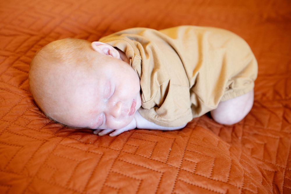 Sleeping newborn baby in a tan outfit curled up on a rust orange quilted blanket.