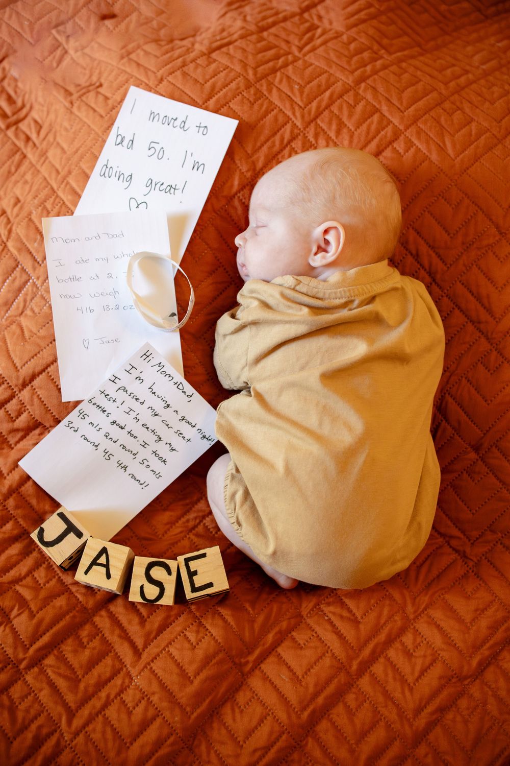Sleeping newborn in a tan outfit on an orange quilted blanket beside handwritten notes, a pacifier, and wooden letter blocks spelling JASE.