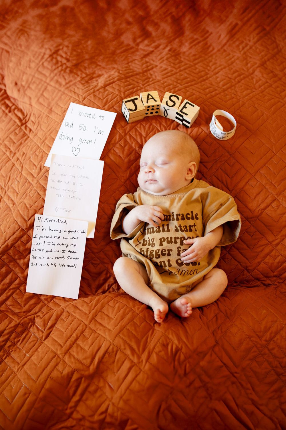 Sleeping newborn baby in a tan graphic onesie on a rust quilted blanket beside handwritten notes, wooden JASE letter blocks and a bracelet.