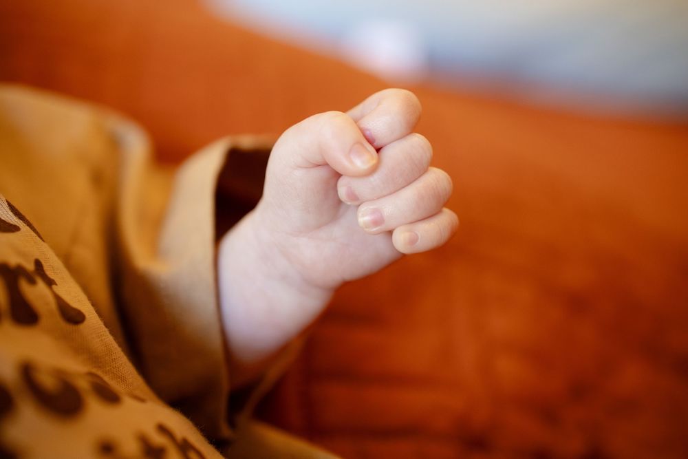 Newborn baby's small hand curled into a gentle fist, resting beside a leopard print blanket on an orange couch.