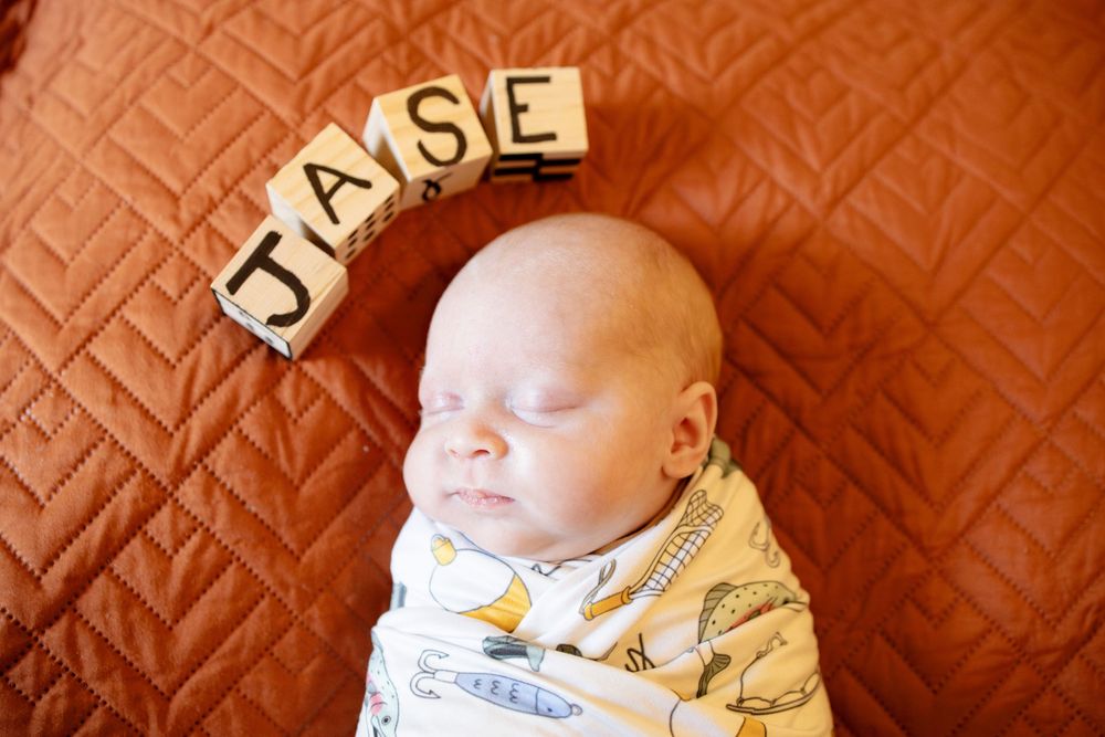 Sleeping newborn in a swaddle lies on an orange quilted blanket beneath wooden letter blocks spelling the name Jase.