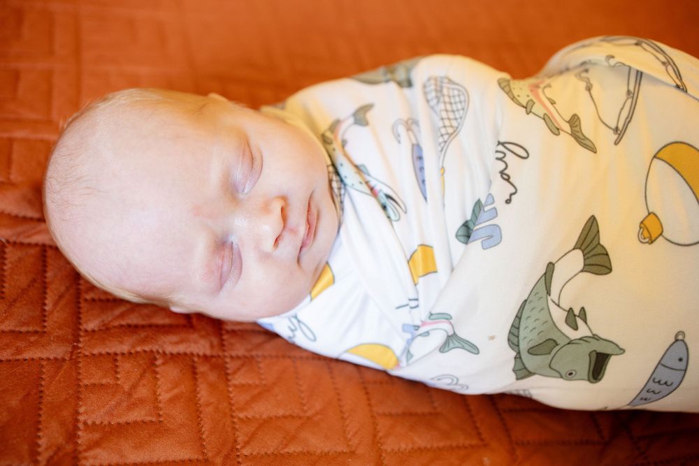 Sleeping newborn baby swaddled in a white blanket with a colorful fish print, lying on a rust orange quilted surface.