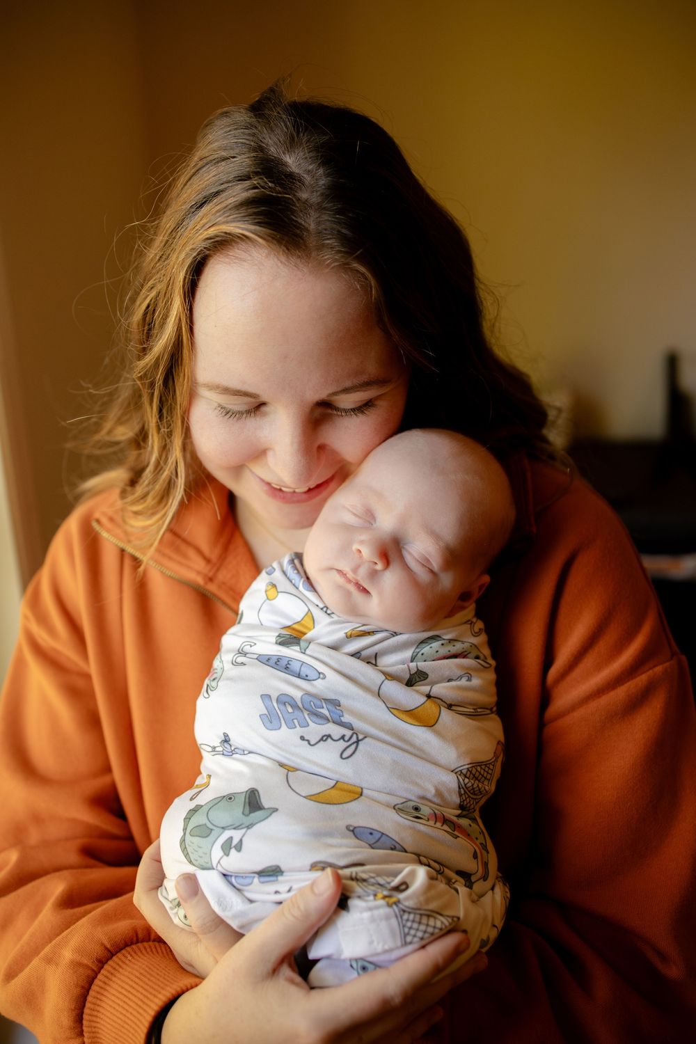 Smiling woman in an orange sweatshirt gently cradles a sleeping newborn baby wrapped in a swaddle printed with the name Jase.