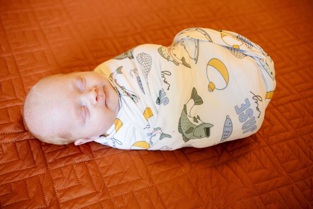 Sleeping newborn baby swaddled in a soft cream blanket with animal and hot air balloon print, lying on a rust orange quilted bedspread.