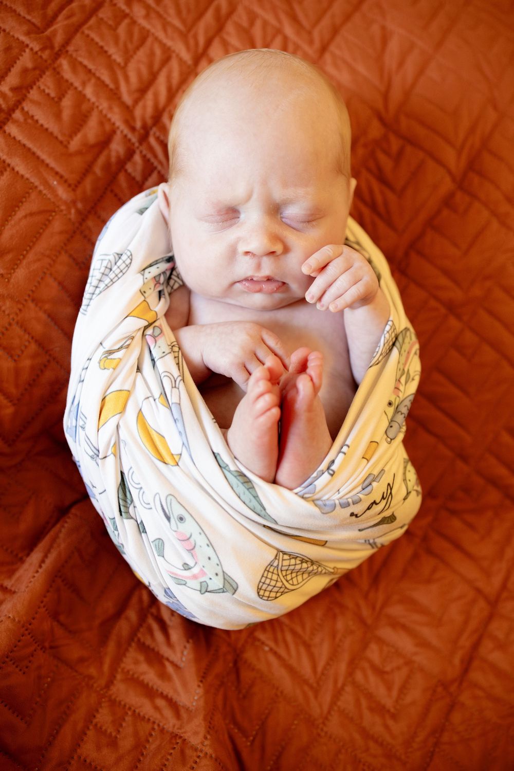 Sleeping newborn baby wrapped in a fish-print swaddle blanket, tiny feet tucked up, resting on an orange quilted background.