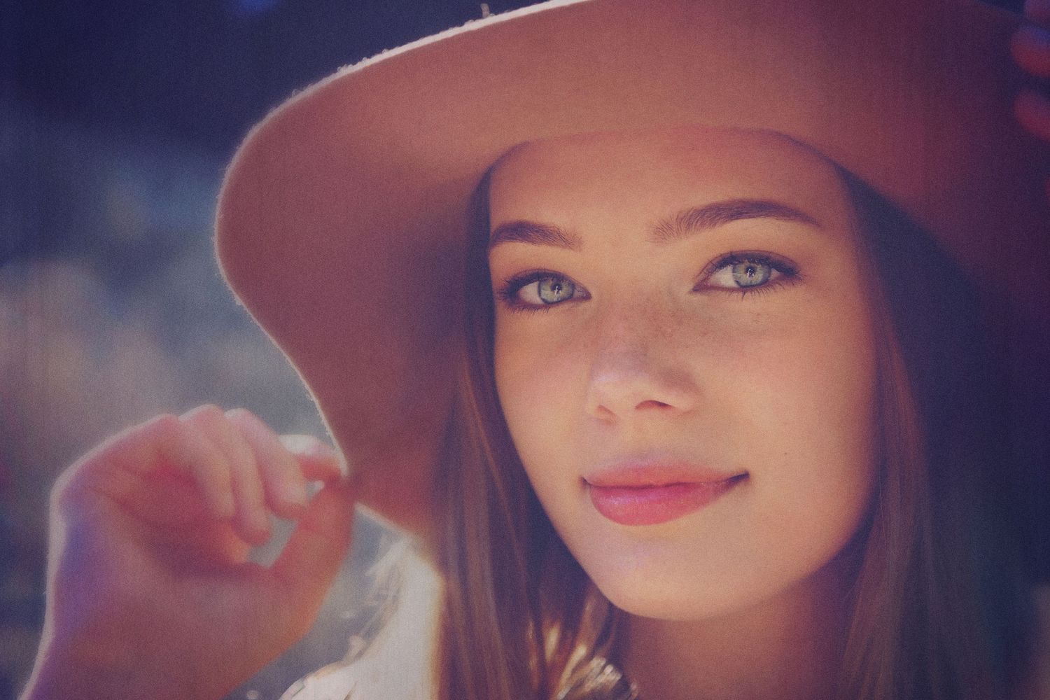 Portrait of a young woman with blue-green eyes and long brown hair wearing a tan hat and touching the brim with her hand.