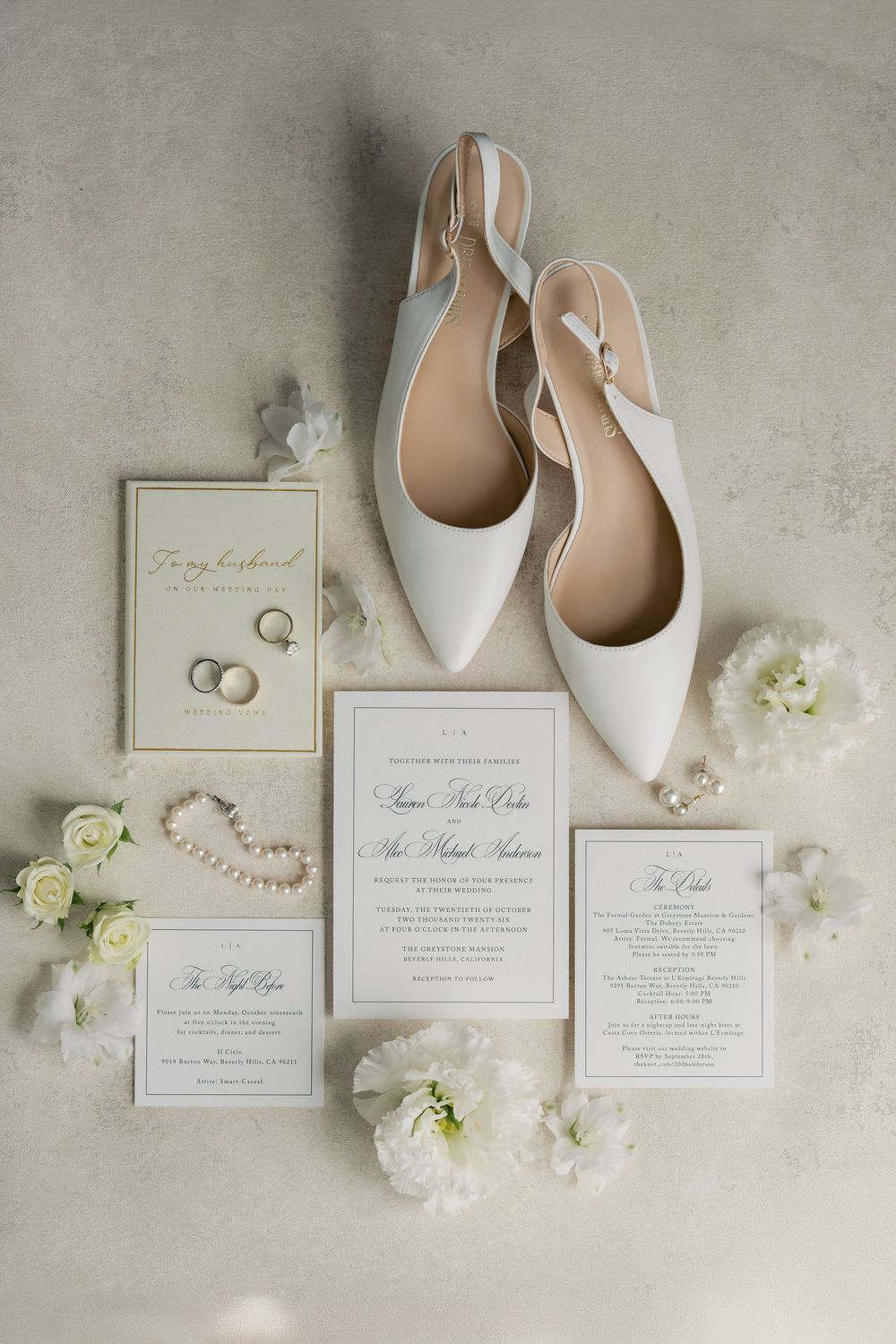 Flat lay of white pointed slingback wedding shoes, pearl bracelet, rings, white flowers and elegant wedding invitation suite.