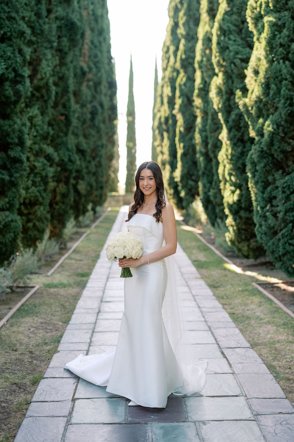 Bride in an elegant white strapless gown holds a white floral bouquet on a stone pathway lined with tall cypress trees.