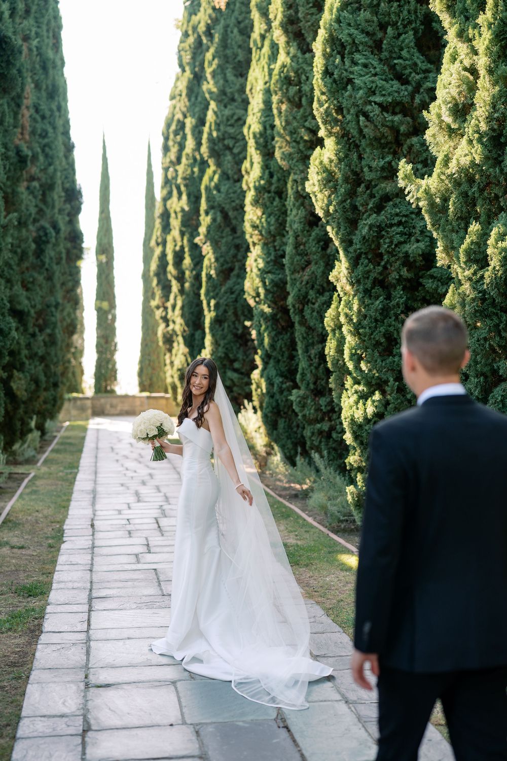 Bride in a white gown and long veil holding a bouquet walks a stone path lined with tall cypress trees toward the groom in a dark suit.