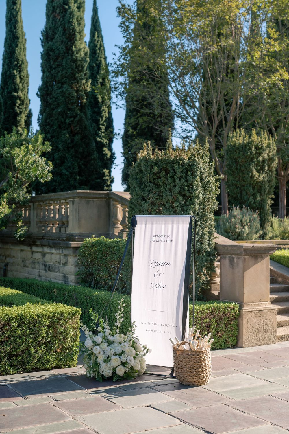 Wedding welcome sign with white floral arrangement and woven basket beside cypress trees and a stone balustrade in a garden.