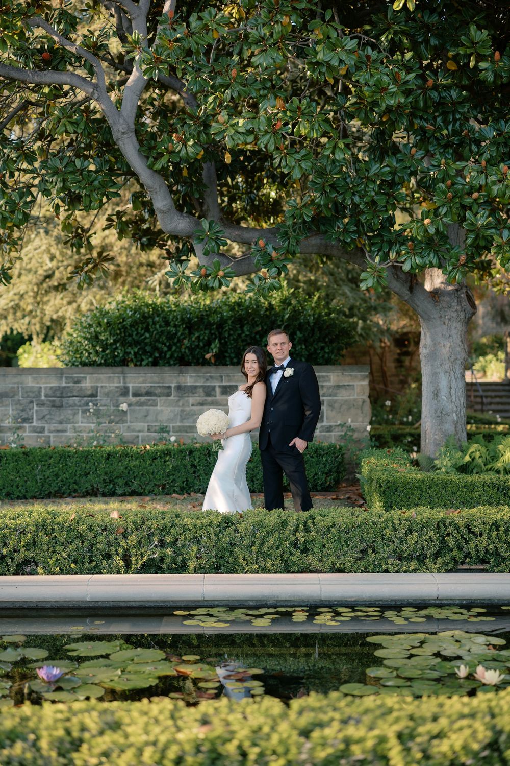 Bride and groom embrace beneath a large magnolia tree beside a reflecting pool with lily pads in a manicured garden.