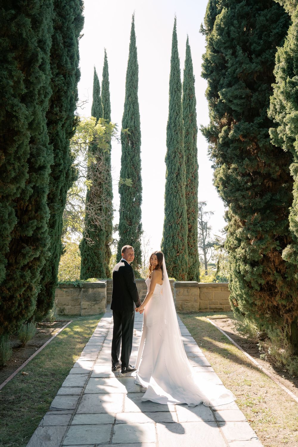Bride and groom hold hands on a stone path lined with tall cypress trees, her long veil trailing behind her.