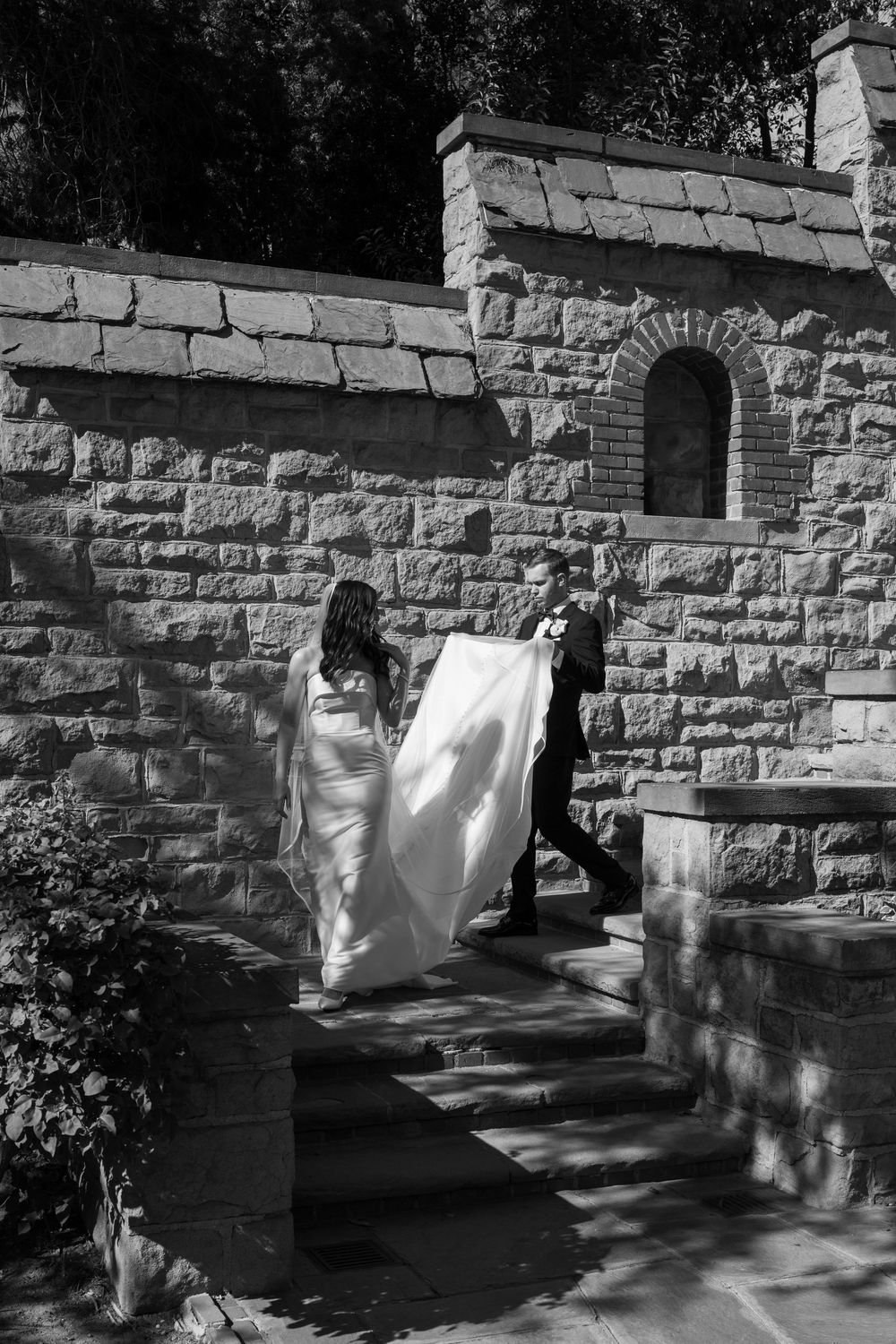 Black and white photo of a groom helping a bride lift her long veil as they walk down stone steps by a brick wall.