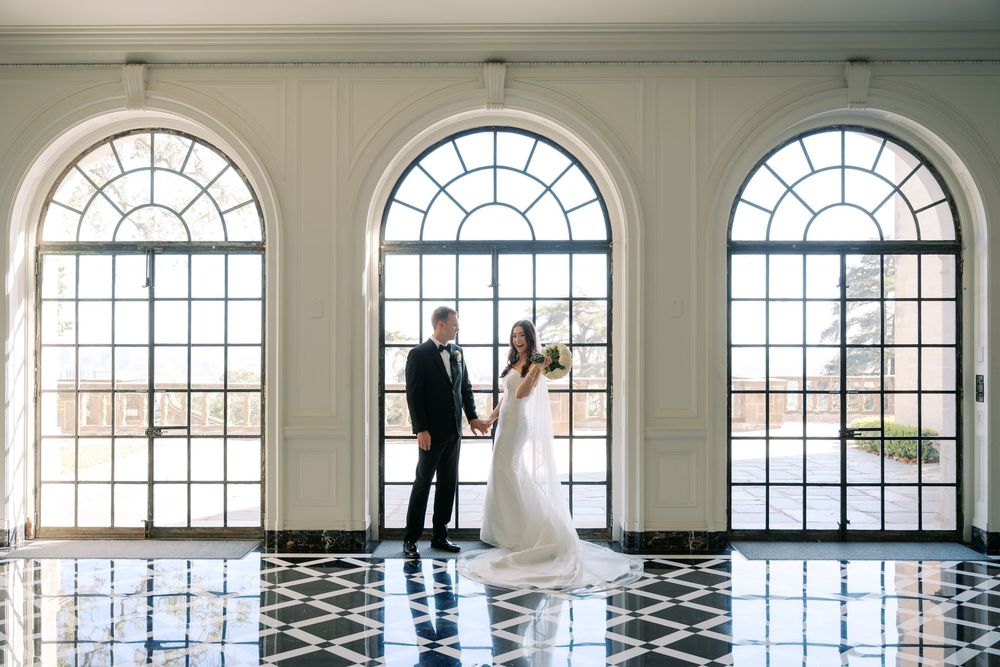 Bride in a white gown and groom in a dark suit hold hands beneath tall arched windows on a black and white marble floor.
