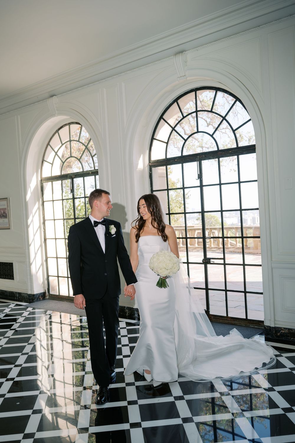 A bride in a strapless white gown holding a bouquet stands hand in hand with a groom in a tuxedo on a black and white checkered floor.