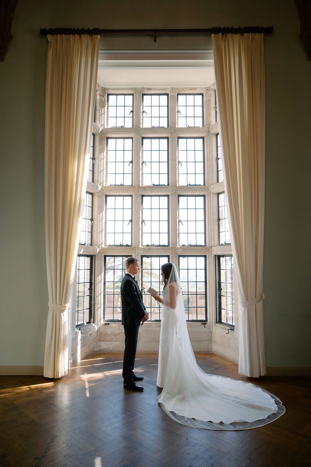 Bride in a long veil and gown faces her groom in front of a tall mullioned window with cream curtains on a wood floor.
