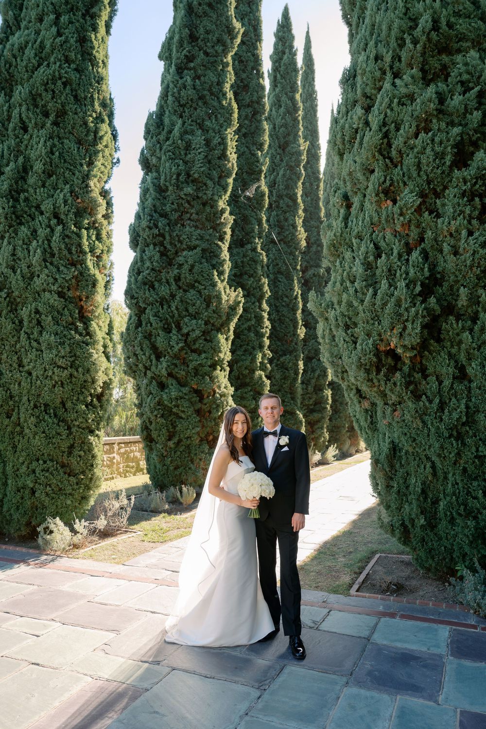 Bride in a white gown holding a bouquet stands beside a groom in a black tuxedo on a stone path lined with tall cypress trees.
