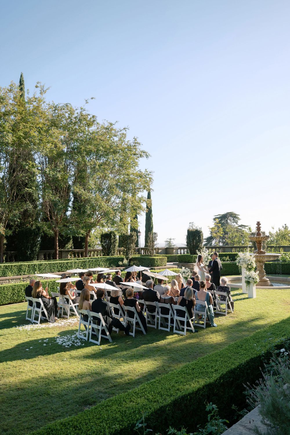 Wedding guests sit in white chairs on a manicured garden lawn as a ceremony takes place near a stone fountain on a sunny day.