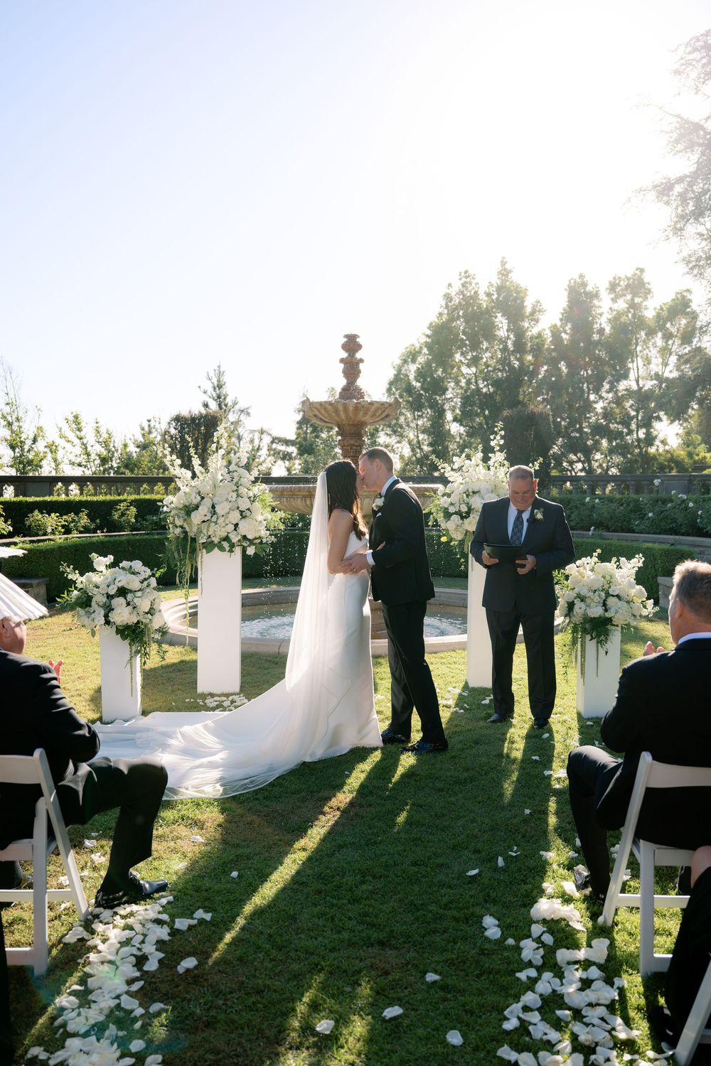 Bride and groom share a kiss at an outdoor garden wedding ceremony with white floral arrangements, a fountain, and guests seated nearby.