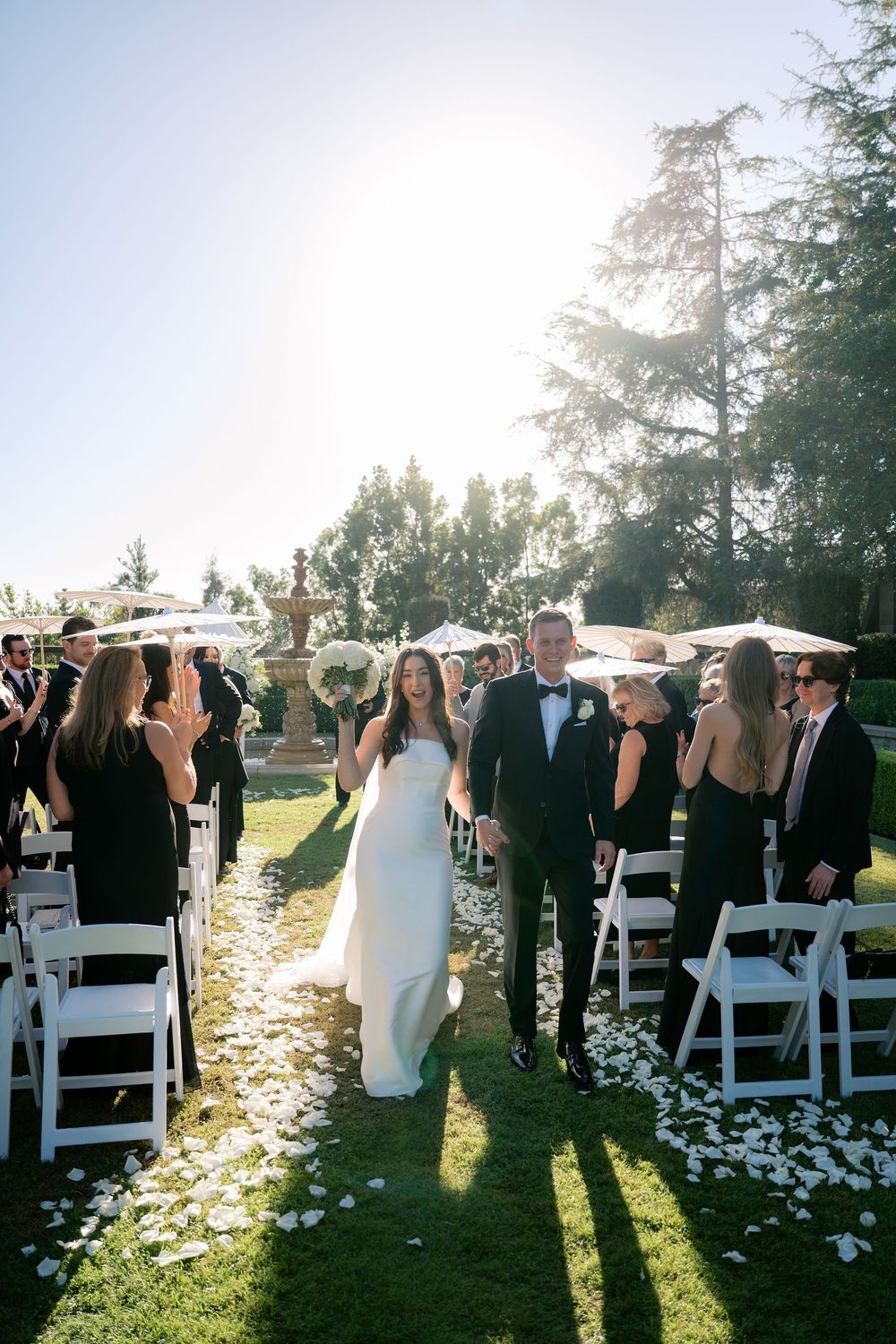 Newlyweds walk hand in hand down a petal-strewn lawn aisle as guests in formal attire cheer at a sunlit garden wedding recessional.