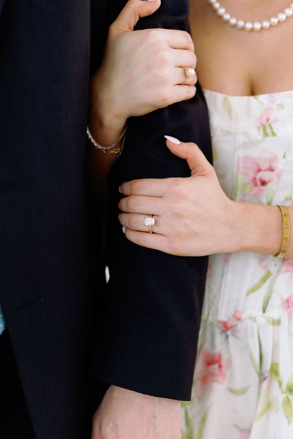 Close-up of woman's engagement ring with diamond solitaire as she holds man's arm in a black suit jacket.
