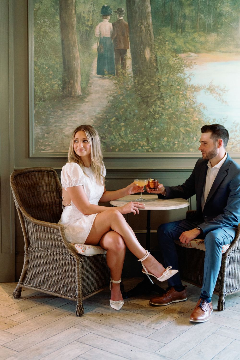 Engaged couple sharing drinks at a small table in an elegant room with a romantic landscape painting backdrop.