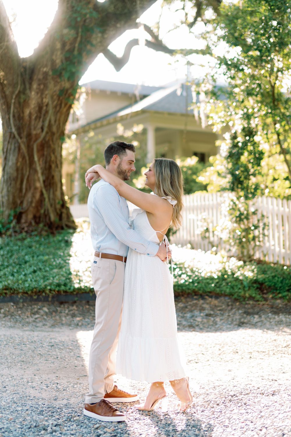 Engaged couple embracing under a large oak tree outside a charming Southern home in golden light.