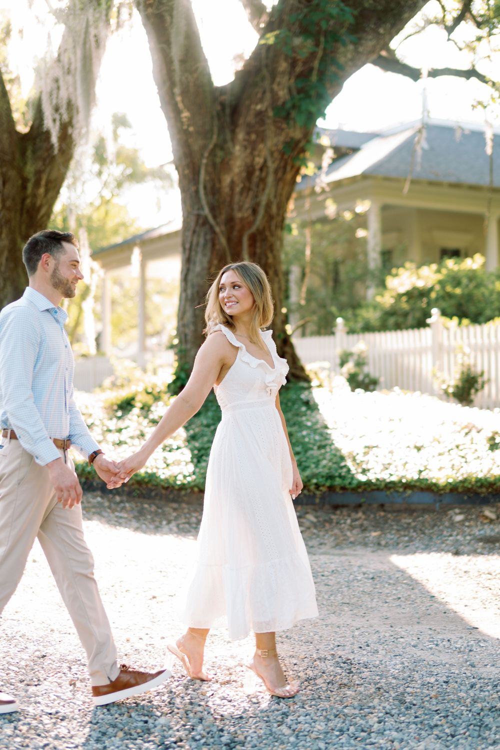 Couple holding hands walking on gravel path under moss-draped oak tree, woman in white midi dress smiling back.
