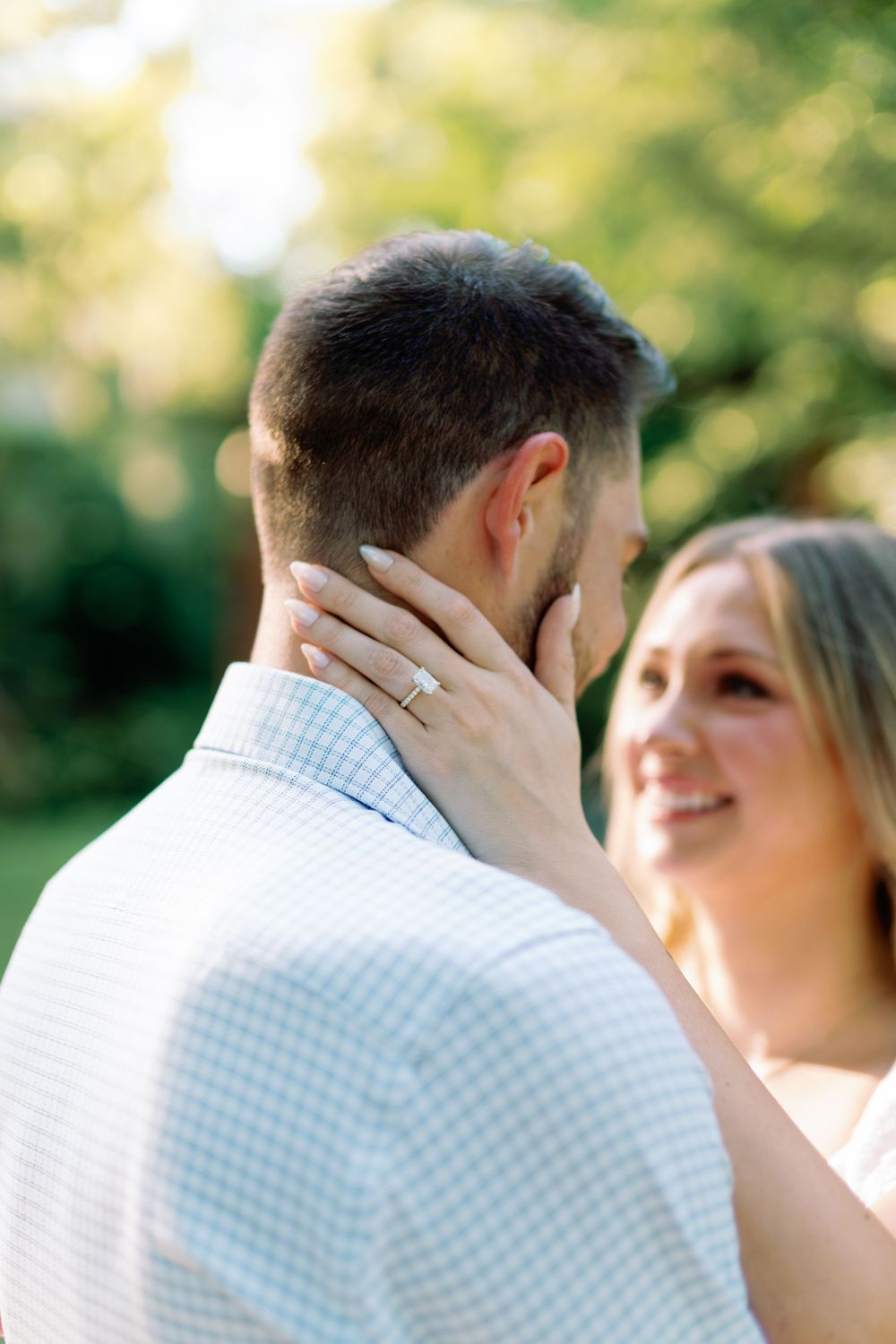 Close-up of woman's hand with diamond engagement ring resting on man's jaw as couple faces each other outdoors.