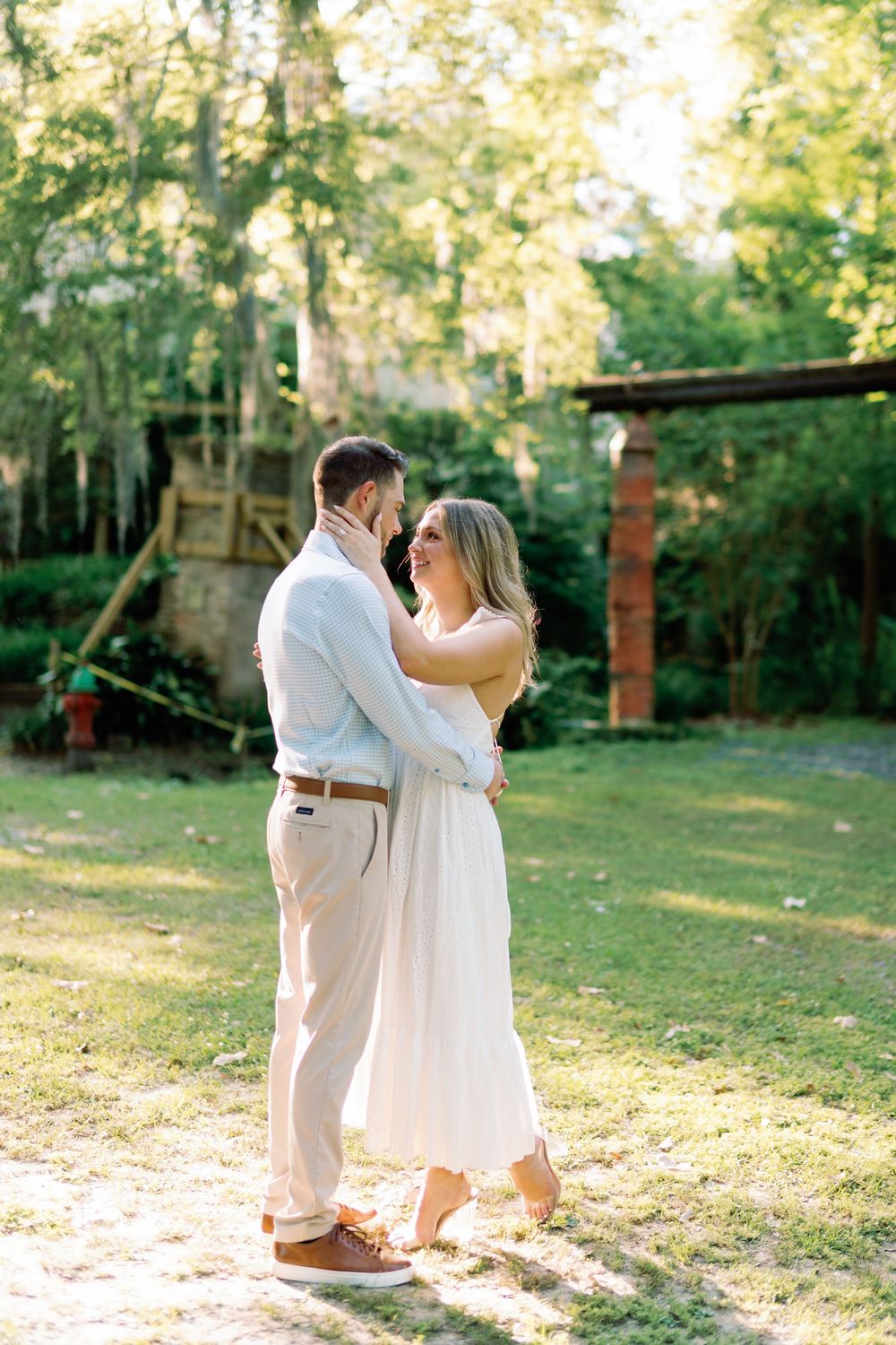 Engaged couple embracing in sunlit garden, woman in white eyelet dress gazing up at man in light blue shirt.