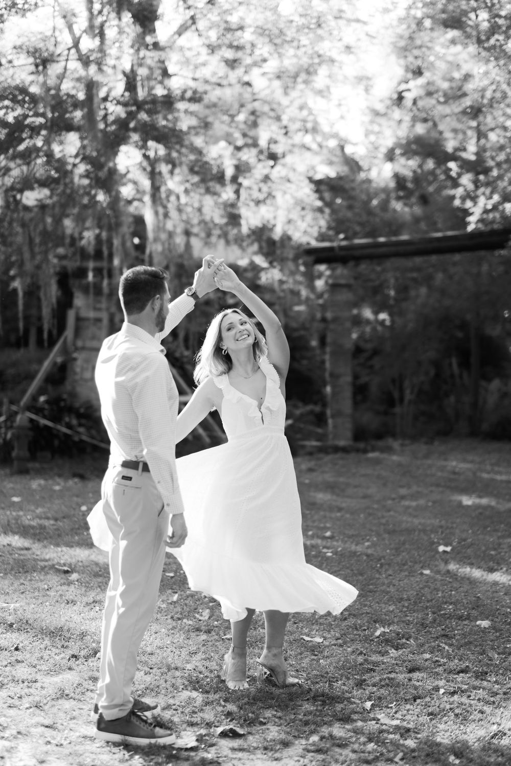 Black and white photo of joyful couple dancing outdoors, man spinning woman in flowing white dress in garden.
