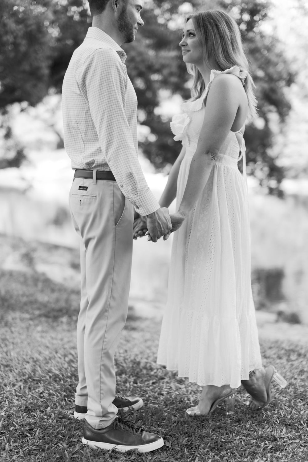 Black and white portrait of engaged couple holding hands facing each other in lush outdoor garden setting.